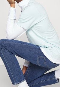 Camisa de manga corta azul claro superpuesta sobre una camisa de manga larga blanca, combinada con jeans azul oscuro que presentan un detalle en el bolsillo trasero.