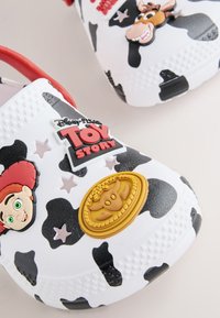 Bele klompe s črnimi kravjimi lisami z liki iz Toy Story Jessie in Bullseye, z zlatim šerifskim značkom in rdečo logotipom Toy Story.