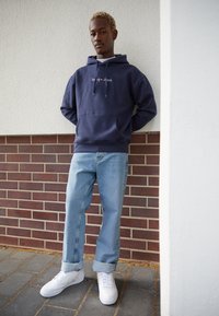 Sudadera con capucha en navy con bolsillo frontal, jeans de denim azul claro y zapatillas blancas. Atuendo casual frente a un fondo de pared de ladrillo.