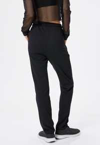 Osoba stojąca tyłem, ubrana w czarne spodnie elastyczne z talią, czarną, siateczkową crop top z długim rękawem oraz czarne sneakersy z białą podeszwą.