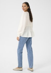 White, long-sleeve blouse with gathered shoulders and tiered hem, paired with light blue jeans and beige shoes.