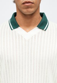 Κοντινό πλάνο ενός ατόμου που φοράει λευκό πουλόβερ πλεκτό με διάτρητο σχέδιο και σκοτεινό πράσινο γιακά με λευκές ρίγες.