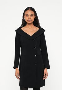 Sieviete ar gariem cirtainiem matiem, valkājot melnu pogātu mēteļa ar platu apkakli un garām piedurknēm, stāv pret balto fonu.