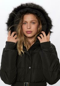 Sorte isolerede pufferjakke med hætte fôret med kunstpels, lynlåslukning og elastiske manchetter. Har en bæltested for et tætsiddende look.