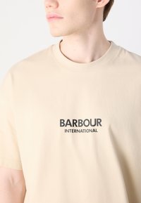Beżowa bawełniana koszulka z okrągłym dekoltem, krótkimi rękawami oraz nadrukowanym logo "BARBOUR INTERNATIONAL" w kolorze czarnym. Gładka faktura.