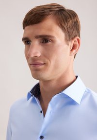 Camicia azzurra chiara con colletto a pois navy in contrasto. Presenta un design aderente e chiusura con bottoni. Tessuto dalla texture liscia.
