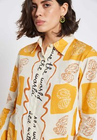Collared shirt in white with yellow and orange abstract oyster prints. Features button closure, long sleeves, and printed text.