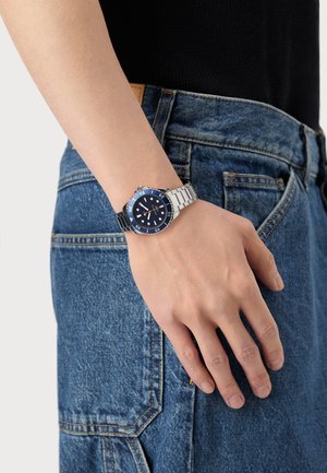 Montre de plongée en acier inoxydable avec un cadran bleu, une lunette argentée et un bracelet en caoutchouc. Main posée sur un tissu en denim, mettant en valeur des coutures détaillées.
