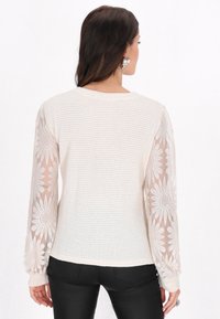 Hellbeiger Pullover mit strukturiertem Oberteil und transparenten langen Ärmeln, die ein florales Muster aufweisen. Gerippte Bündchen und runder Ausschnitt.