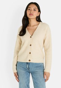 Cardigan de couleur crème en matériau doux, avec un col en V, cinq boutons marron sur le devant, et des poignets et un ourlet côtelés.
