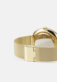 Złota siateczkowa bransoletka zegarka z zapięciem z logo "Calvin Klein". Okrągła złota obudowa z szklanym tyłem, podkreślająca elegancki design i fakturę.
