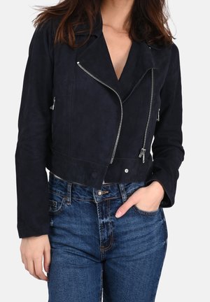 Frau trägt eine schwarze, kurz geschnittene Bikerjacke mit silbernen Reißverschlüssen und blaue, hoch taillierte Jeans, die Hand in die Tasche gesteckt.