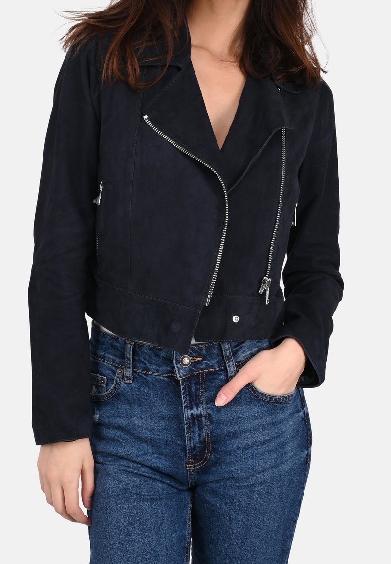 Kvinde iført en sort croppet bikerjakke med sølv lynlåse og blå højtaljede jeans, hånd stukket i lommen.