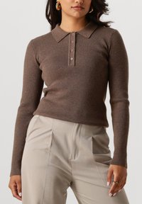 Frau in einem enganliegenden braunen Ripp-Polo-Pullover mit langen Ärmeln und hoch taillierten beigen, gefalteten Hosen, die ihren Oberkörper und ihre Hände zeigt.