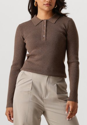 Vrouw in een getailleerde bruin geribbelde polo trui met lange mouwen en high-waisted beige geplooide broeken, waarbij ze het middel en haar handen toont.