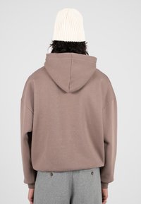 Persona che indossa una felpa con cappuccio taupe, pantaloni grigio chiaro con bottoni e un berretto di maglia color crema, vista da dietro su uno sfondo semplice.