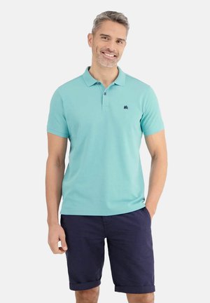 Mann lächelt, trägt ein hellblaues Poloshirt und dunkelblaue Shorts, steht mit einer Hand in der Tasche vor einem schlichten weißen Hintergrund.