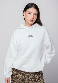 Sudadera blanca con un bolsillo frontal y el texto "ANARQUISMO FELINO". Combinada con pantalones de estampado de leopardo. Tejido suave, corte holgado y capucha grande.