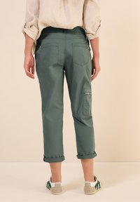 Gröna bomullsbukser med uppvikta cuffs, bakfickor och en sidoficka med dragkedja. Bärs med grå och vita sneakers. Neutral bakgrund.