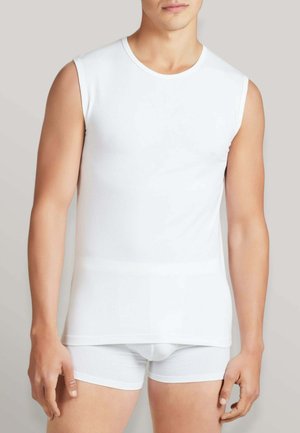 Uomo che indossa una canottiera bianca semplice senza maniche e boxer bianchi abbinati, in piedi davanti a uno sfondo grigio chiaro.