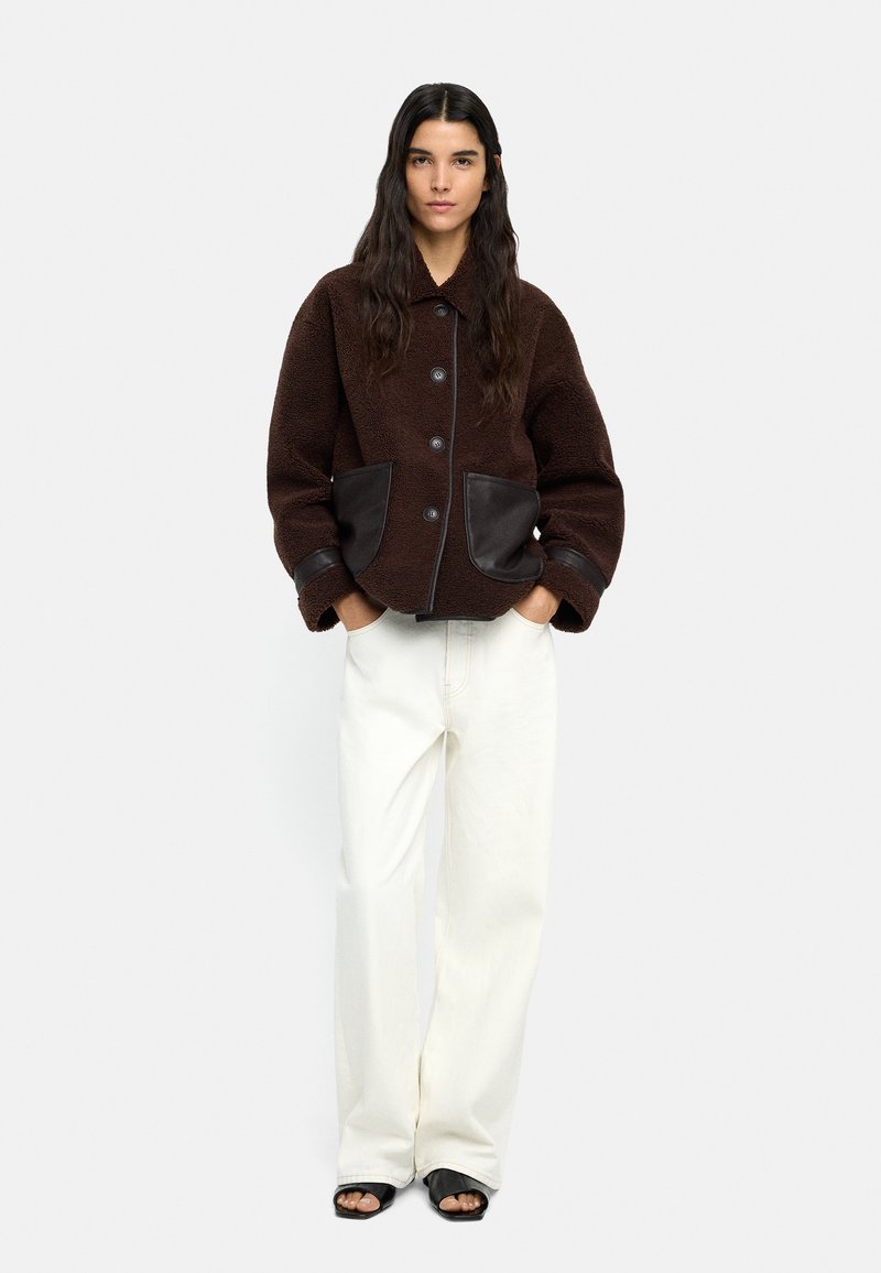 Brązowa bluza polarowa z czarnymi skórzanymi akcentami i kieszeniami, połączona z białymi spodniami o szerokich nogawkach oraz czarnymi klapkami.