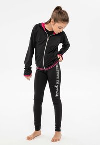 Giacca atletica nera con dettagli rosa, con zip sul davanti e maniche lunghe. Leggings neri con la scritta bianca "Credi in te stesso" lungo il lato.