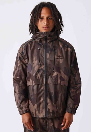 Jeune homme aux cheveux mi-longs bouclés portant une veste à capuche camouflage marron et noir, debout devant un fond clair uni.