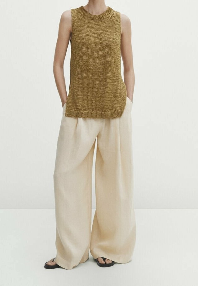 Haut marron sans manches en tricot associé à un pantalon beige large et ample, et des sandales noires, modèle debout les mains dans les poches.