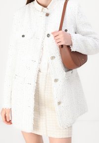 Witte tweed jasje met rafelige randen, gouden en zwarte knoopaccenten, gecombineerd met een lichte blouse en een beige gestructureerde rok. Bruine schoudertas.