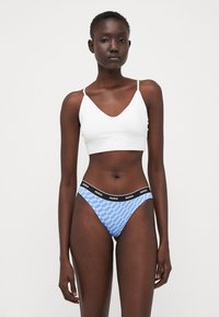 White crop top with thin straps paired with blue thong featuring black HUGO logo waistband. Smooth fabric, minimalist design, fitted style.