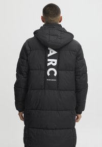 Pufferjakke i svart, med hette, quiltet tekstur og en vertikal hvit logo på baksiden: "ARC SOLID ARCHIVE." Lang lengde.