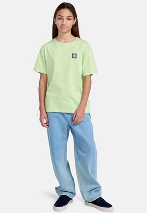 Mint green cotton t-shirt with a round neck and short sleeves, featuring a small black logo patch. Worn with light blue baggy jeans.