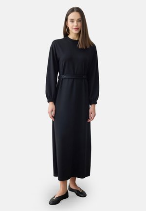Kvinde iført en lang sort kjole med bælte i taljen og lange ærmer, sorte flade sko og gyldne hoops-øreringe, stående mod en ensfarvet baggrund.