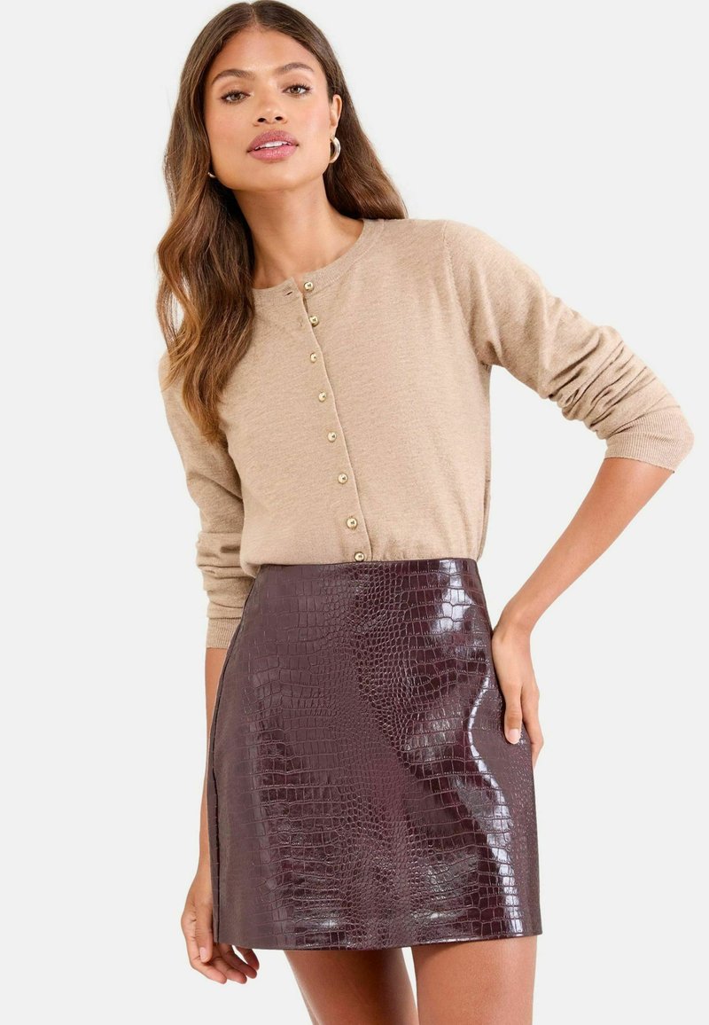 Maglione beige a maniche lunghe con bottoni dorati abbinato a una mini gonna in faux-coccodrillo bordeaux, caratterizzata da una trama liscia e lucida.