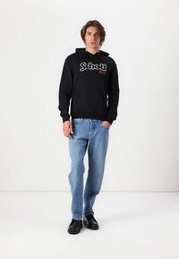 Sweat à capuche noir avec logo "Schott NYC" en blanc et rouge, porté avec un jean bleu clair et des chaussures noires, doté d'une capuche à cordon.