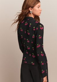 Chemise noire à manches longues présentant une texture côtelée et un motif de cerises en rose et vert. Coupe ajustée avec un col et des détails subtils.