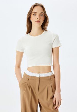 Witte cropped t-shirt van ribbelstof, met ronde halslijn, korte mouwen, gecombineerd met bruine plooibroek met een elastische tailleband.