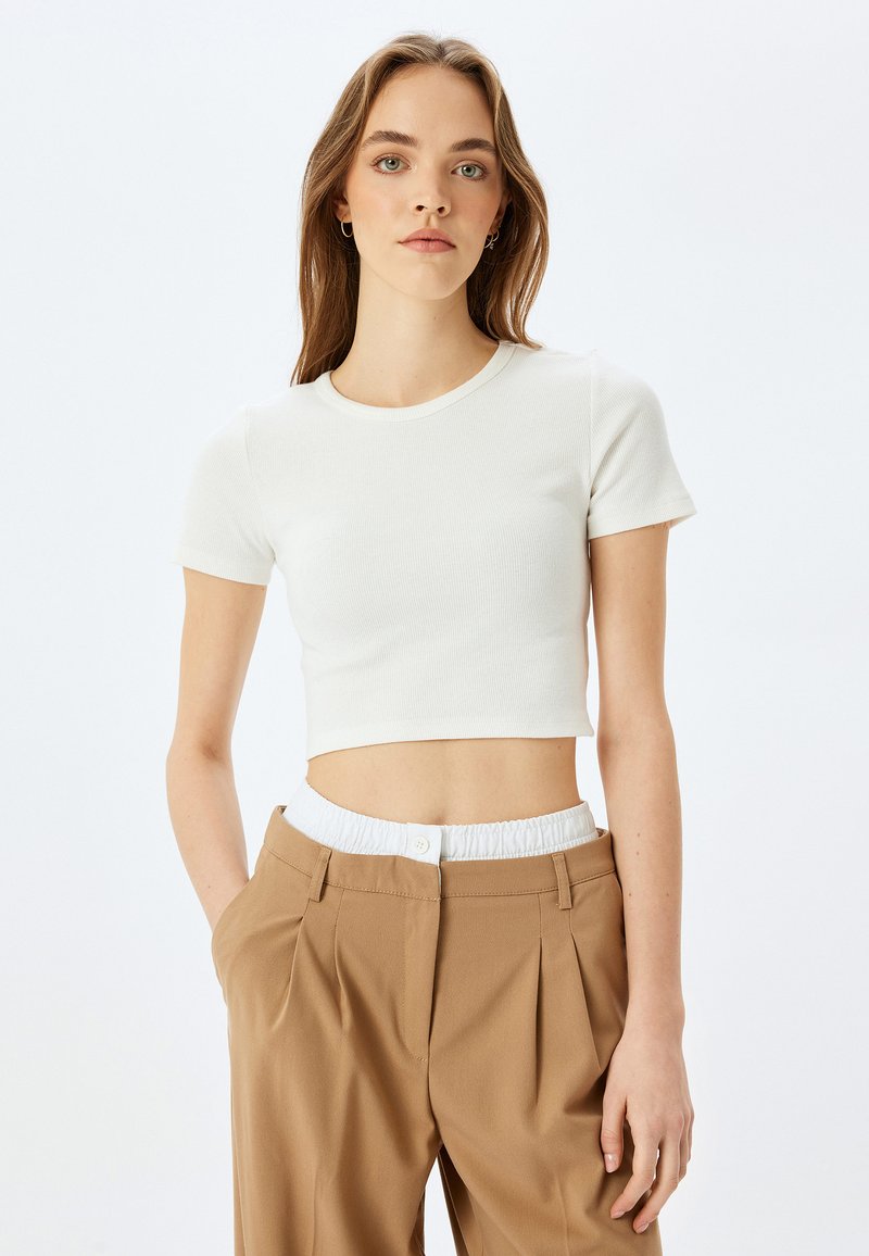 Hvid cropped t-shirt i ribbet stof, rund halsudskæring, korte ærmer, parret med brune plisserede bukser med elastisk taljebånd.