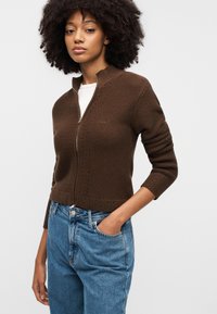 Καφέ πλεκτό πουλόβερ με φερμουάρ και ψηλό γιακά, με υφασμένη υφή, συνδυασμένο με ανοιχτό μπλε ψηλόμεσα τζιν.