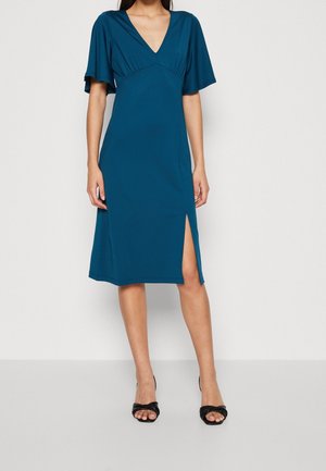 Vrouw draagt een teal knielange jurk met vliegende mouwen en een elegante zijsplit, gecombineerd met zwarte open-teehakken tegen een effen achtergrond.