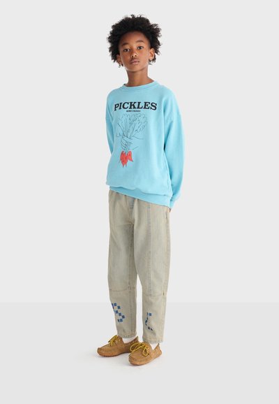 Bambino in piedi che indossa una felpa azzurro chiaro con la scritta "PICKLES" e verdure disegnate a mano, pantaloni beige con toppe blu e scarpe marroni.