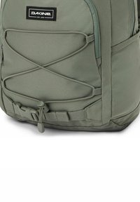 Mochila de tela verde con un diseño de cuerda elástica diagonal, cierre de hebilla y un bolsillo lateral de malla para almacenamiento adicional.