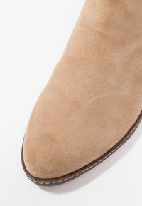 Chaussure en daim beige à bout pointu avec couture de la semelle marron sur fond blanc, vue en angle depuis un coin supérieur.