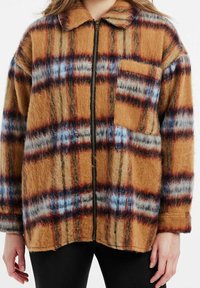 Veste zippée à carreaux marron avec des accents bleus et rouges, fabriquée en matière duveteuse. Comprend un col et une poche poitrine unique.