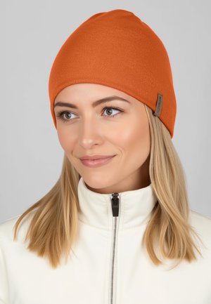 Oranje gebreide muts met een aansluitende pasvorm, gemaakt van zacht materiaal. Heeft een klein bruin label aan de zijkant. Draagt een witte jas met rits.