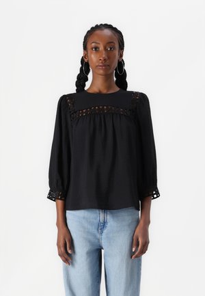 Schwarze Bluse mit quadratischem Ausschnitt, strukturiertem Stoff und Dreiviertelärmeln. Verfügt über ausgeschnittene Spitzenakzente an den Schultern und Manschetten.