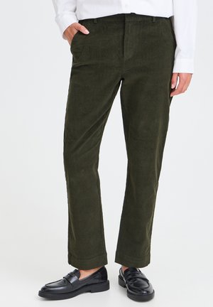 Persona que lleva pantalones de pana verde oscuro, camisa blanca de manga larga y mocasines de cuero negro, de pie con una mano en el bolsillo.