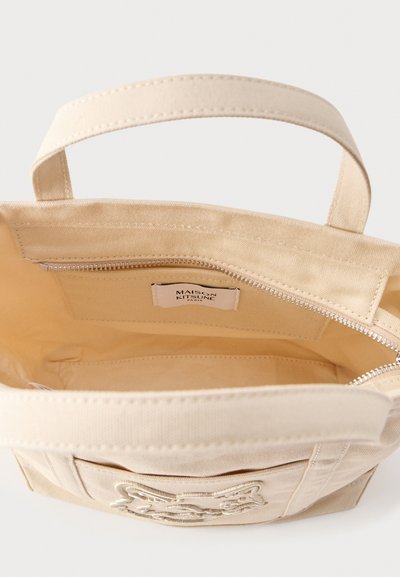 Μπεζ τσάντα ώμου από καμβά με δύο λαβές, κεντητό σχέδιο αλεπούς στο μπροστινό μέρος, εσωτερική τσέπη με φερμουάρ και ετικέτα Maison Kitsuné μέσα.