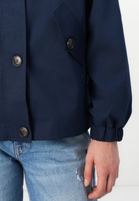 Marineblå jakke med sorte knapper, en klaffelomme og rynkede mansjetter. Laget av glatt stoff, har en avslappet passform og rene linjer.