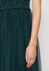 Rochie verde închis cu un corset din dantelă și o fustă din tull stratificată. Țesătura are o finisare texturată, cu detalii delicate la talie.
